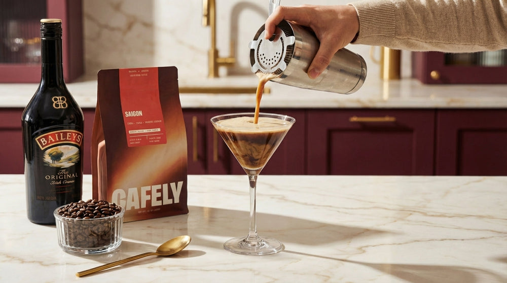 Coffee cocktail being poured into a martini glass from a shaker, beside Baileys, coffee beans, and a Cafely Saigon coffee bag.
