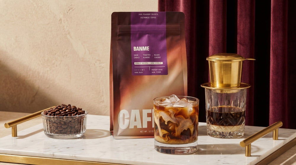 Iced coffee with cream swirling in a glass beside a Vietnamese phin brewer, coffee beans, and a Cafely BanMe coffee bag.