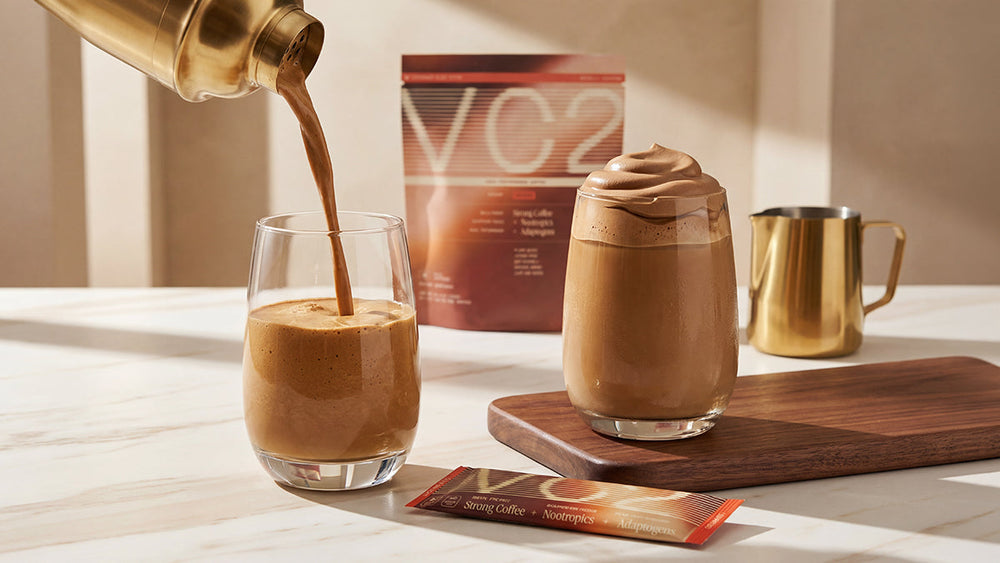 Coffee frappe being poured into a glass beside a whipped coffee drink, with a VC2 coffee bag and shaker.