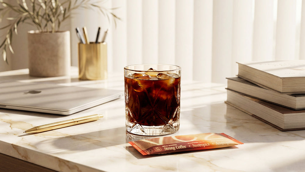 Glass of iced coffee on a desk with books, a laptop, and a stick pack of instant coffee nearby.